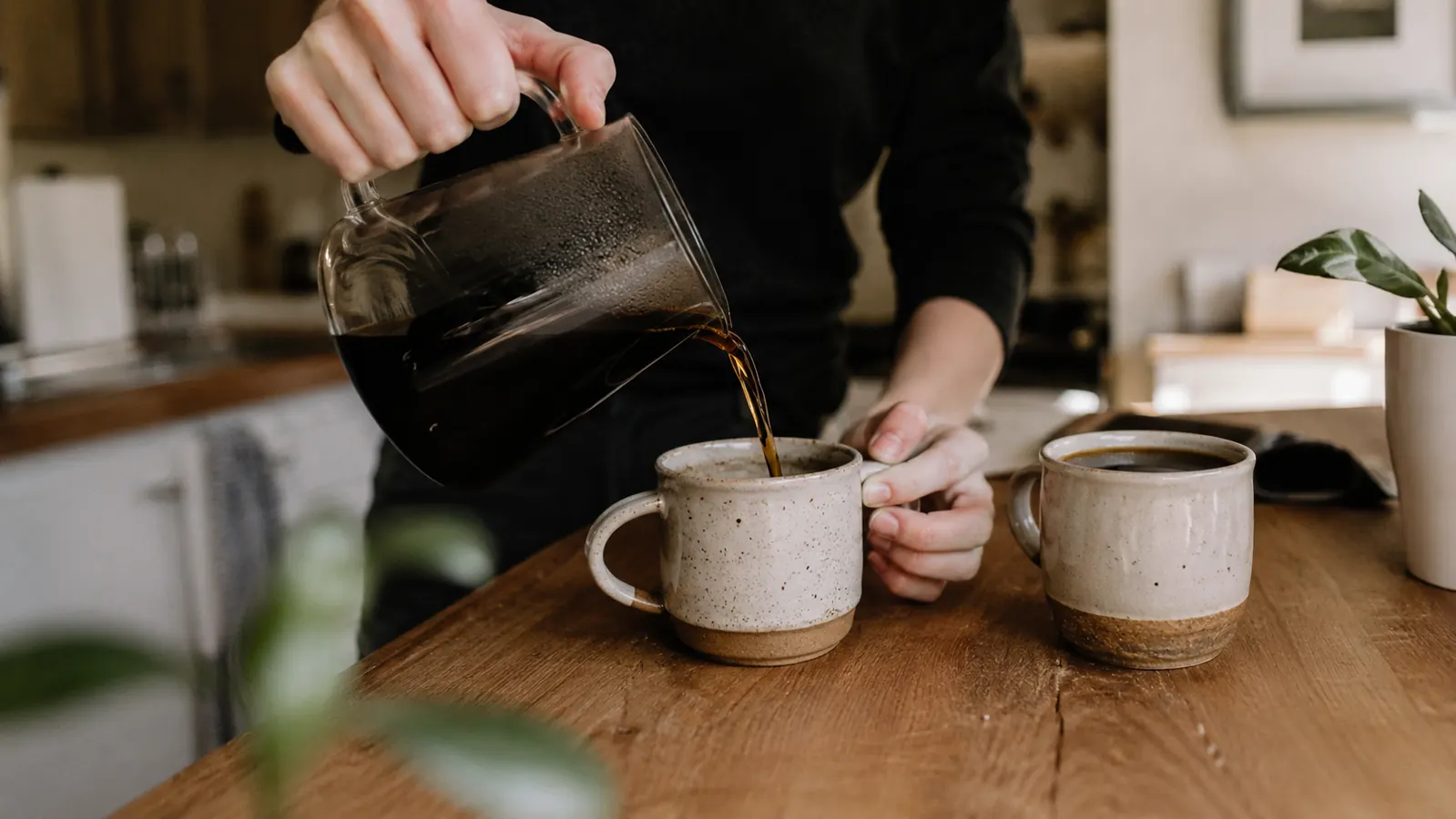 A full glass carafe of freshly brewed coffee being poured into a mug on a small kitchen counter, second cup visible in the background