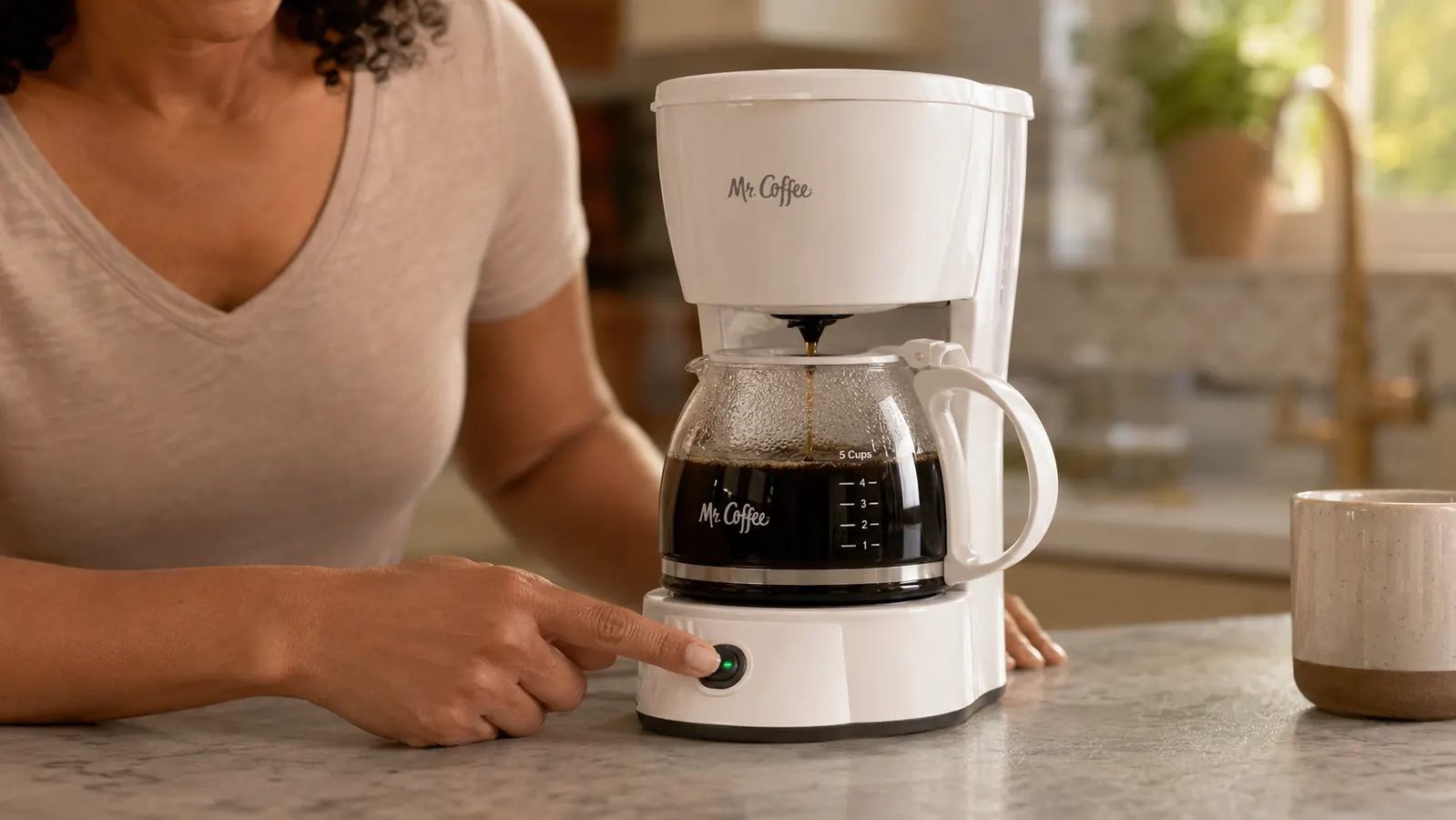 Hand pressing the brew button on the Mr. Coffee 5-Cup Mini while the glass carafe fills with fresh coffee